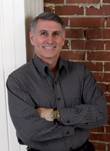Dr. Bill Morgan leans against a brick wall, smiling, with his arms crossed.
