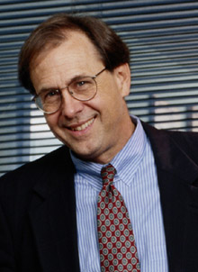 A businessman wearing a suit, tie, and eyeglasses smiles.